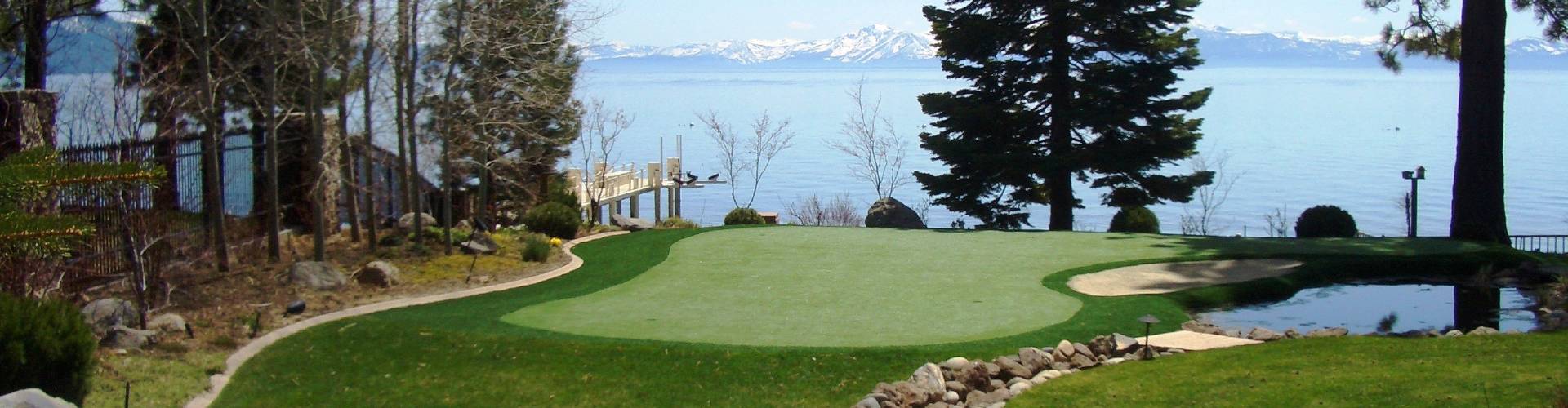 Southwest Greens putting green with sand bunker overlooking a lakeside dock, pine trees, and snow-capped mountain range.